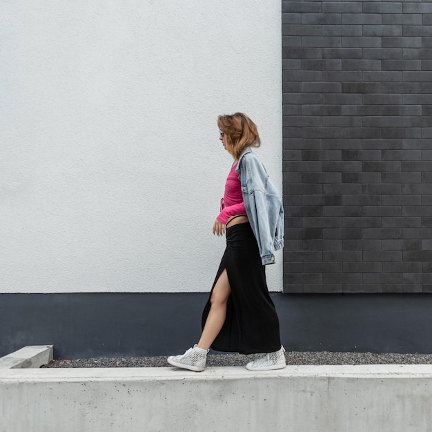 Stylish beautiful young redhead model woman with white sunglasses in fashionable street clothes with a pink sweatshirt long skirt jeans jacket and sneakers walking near a white and black brick wall