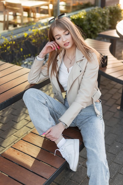 Stylish beautiful young woman hipster in fashionable rock streetwear with leather jacket top blue vintage jeans and white sneakers sits on wooden bench near a cafe