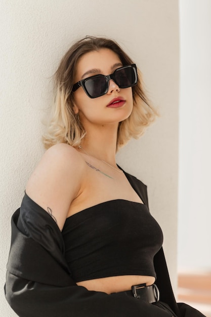 Stylish beautiful young woman with blonde short hair and cool sunglasses in fashion elegant clothes with a top and blazer on the street near the white wall Urban female style look