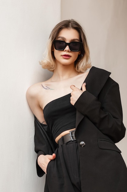 Stylish beautiful young woman with trendy modern sunglasses in fashionable black business clothes with top fashionable blazer and pants standing near the white wall on the street Female urban style