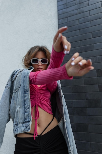 Stylish fashion beautiful redheaded woman with cool white sunglasses in a trendy pink sweatshirt top with jeans jacket and skirt dancing on the street near a black white brick wall