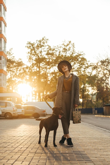 Stylish lady in a coat and hat walking a dog on a leash in the autumn evening at sunset