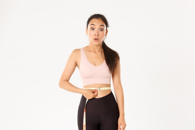 Photo  surprised asian girl on diet, sportswoman wrap measuring tape around waist and look impressed as lose weight with workout.