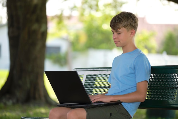 Teenager school boy typing on laptop computer sitting on bench outdoors on summer sunny day Education during quarantine concept