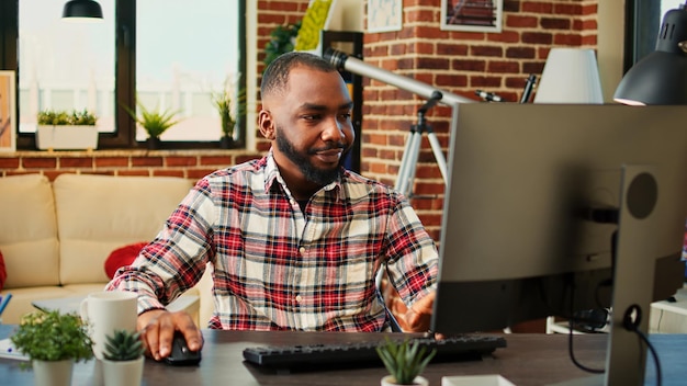 Tired sleepy african american employee remotely working, typing data on computer, happy about finishing work soon. Teleworker answering job emails in warm cozy living room apartment