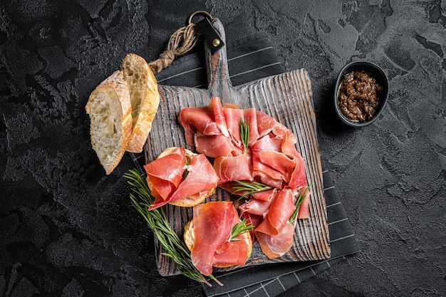 Toast with tomatoes and cured Slices of jamon serrano ham prosciutto crudo parma on wooden board with rosemary Black background Top view