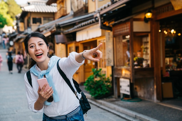 Photo 日本の伝統的な通りに立ち、楽しそうに店を指差す観光客。日本旅行中にスマートフォンでオンラインガイドを使用する女性バックパッカー。アジアのコンセプトでの独立した旅行。