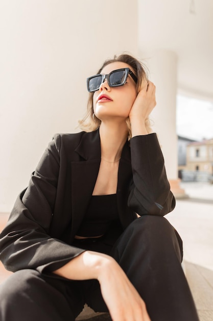 Trendy business fashion beauty woman model with black stylish sunglasses in black urban elegant outfit with top blazer and pants sits in the city near a white vintage column