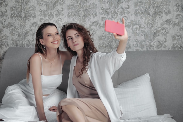 Two adult women in stylish casual white clothes take selfie on bed, chat sweetly, tell stories and laugh. Friends met and spend their leisure time in hotel room. Concept of friendship