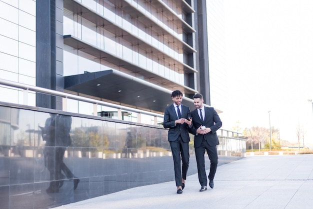 Two businessmen wearing suits walking and using phone smiling and talking outdoor in a business area