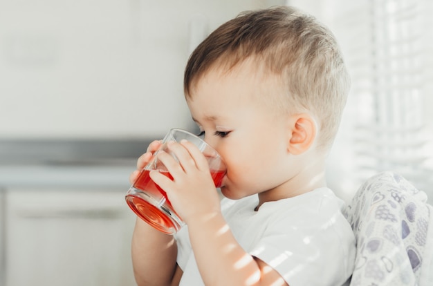 Two years boy drinking juice from a glass Cup in the kitchen