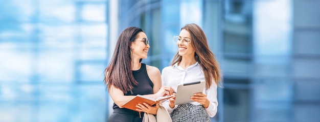 Two young business lady with tablet and notebook walking outdoors near modern business center