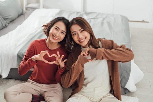 Photo two young women in bedroom at home lying on bed making heart shape with hands