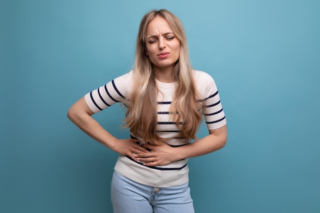 Unhappy girl in casual clothes clutches her stomach due to indigestion on a blue isolated background