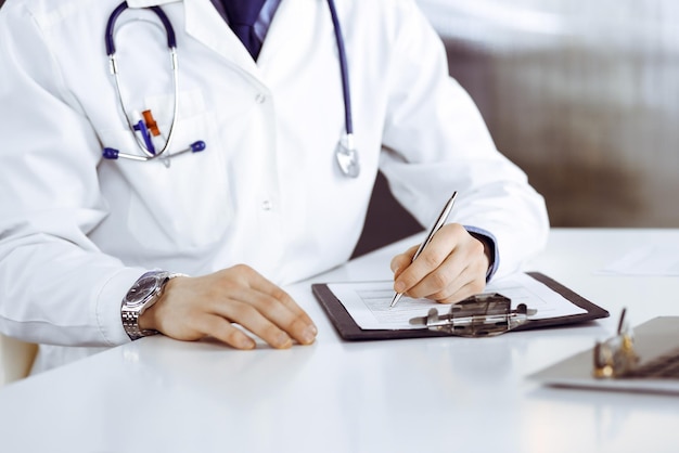 Unknown male doctor sitting and working with clipboard of medication history record in clinic at his working place, close-up. Young physician at work. Perfect medical service, medicine concept.