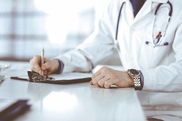 Unknown male doctor sitting and working with clipboard of medication history record in clinic at his working place closeup Young physician at work Perfect medical service medicine concept