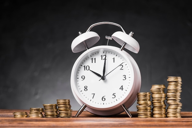 White alarm clock between the increasing stack of coins on wooden table against gray background