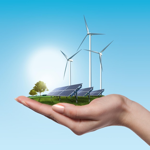 Wind turbines and solar panels on meadow with tree holds in woman's hand against blue sky and clouds