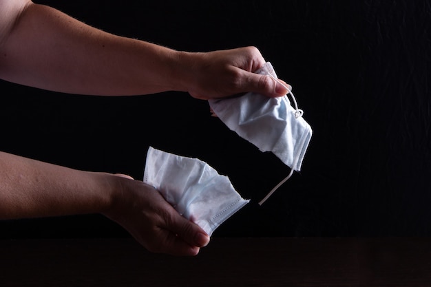 Women's hands tear the medical mask. 2 parts of mask are in hand. Low key.