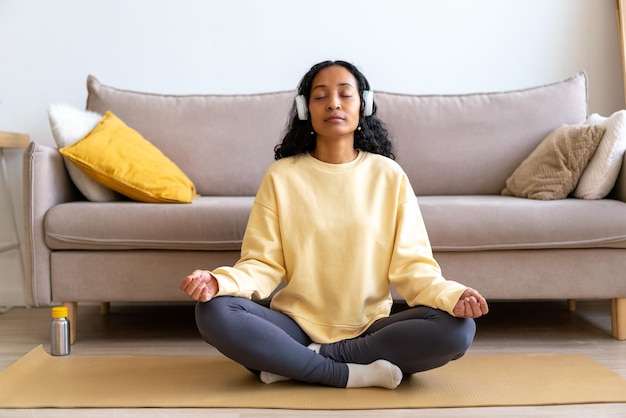 Young africanamerican female in headphone listening to music while meditating on mat in living room