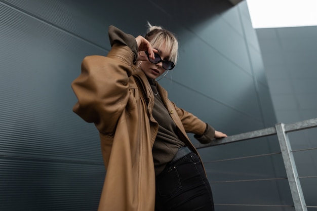 Young beautiful model woman with fashionable sunglasses in brown leather jacket with black jeans posing in the modern city