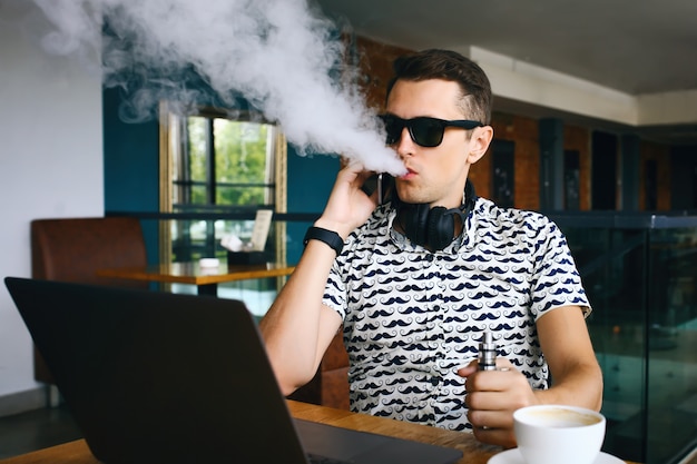 Photo young handsome hipster man insunglasse sitting in cafe with a cup of coffee, vaping and releases a cloud of vapor.