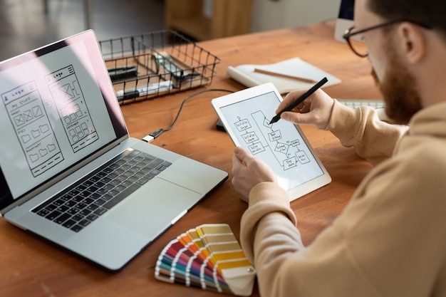 Photo young male freelance software developer holding stylus over tablet screen while drawing sketch of scheme of new smartphone models