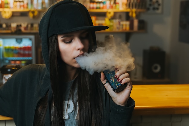 Photo young pretty woman in cap smoke an electronic cigarette at the vape shop. hip-hop style. closeup.