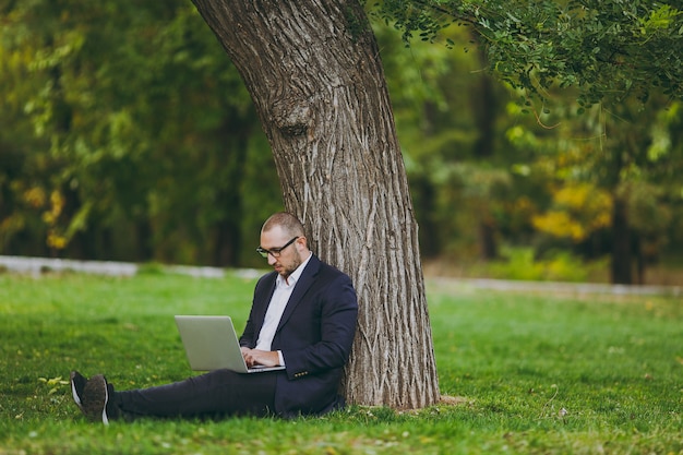 Young successful businessman in white shirt, classic suit, glasses. Man sit on grass ground, work on laptop pc computer in city park on green lawn outdoors on nature. Mobile Office, business concept.