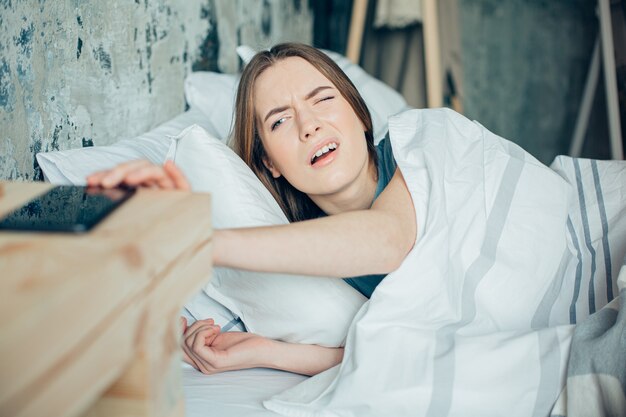 Young woman lying in bed and opening one eye to get her ringing phone in the morning