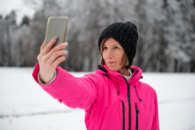 Young woman in pink winter jacket and black hat taking a selfie with her phone standing in beautiful snowy nature.