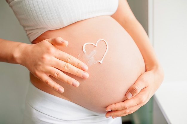 Photo young woman's hand holding a white heart shape made of cream on a naked belly. close-up.