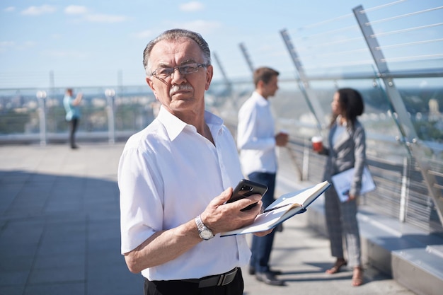 Dojrzały biznesmen ze smartfonem i notatnikiem stojący na tarasie centrum biznesowego