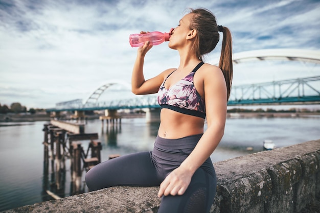 Młoda kobieta fitness odpoczywa po ciężkim treningu na ścianie przy moście nad rzeką i wodą pitną.