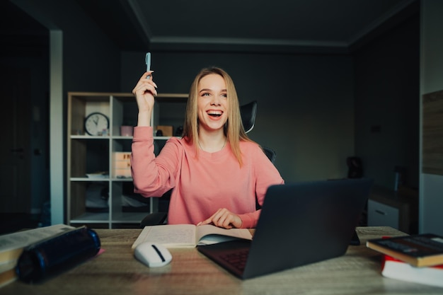 Radosny młody student studiuje w domu i ma pomysł siada przy stoliku obok laptopa