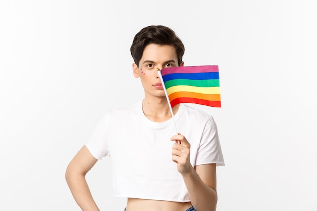 Społeczność Lgbtq. Atrakcyjny queer mężczyzna z falowaniem na twarzy, machając tęczową flagą dumy i patrząc na kamery, stojąc w crop top przed białym.