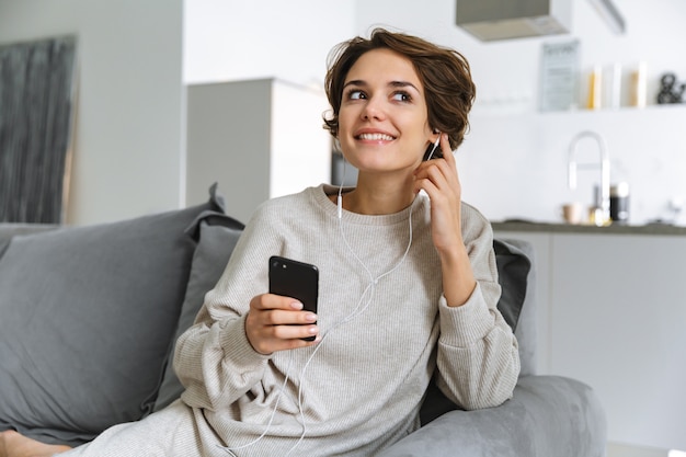 Szczęśliwa młoda kobieta siedzi na kanapie w domu przy użyciu telefonu komórkowego, słuchając muzyki przez słuchawki