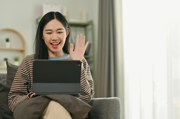 Zdjęcie wesoła młoda azjatka uśmiecha się i macha ręką do cyfrowego tabletu podczas rozmowy wideo