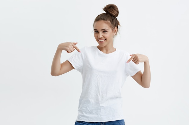 Gratis Foto charmante emotionele blanke vrouw die naar beneden of naar haar t-shirt wijst terwijl ze vrolijk lacht en positieve emoties uitdrukt