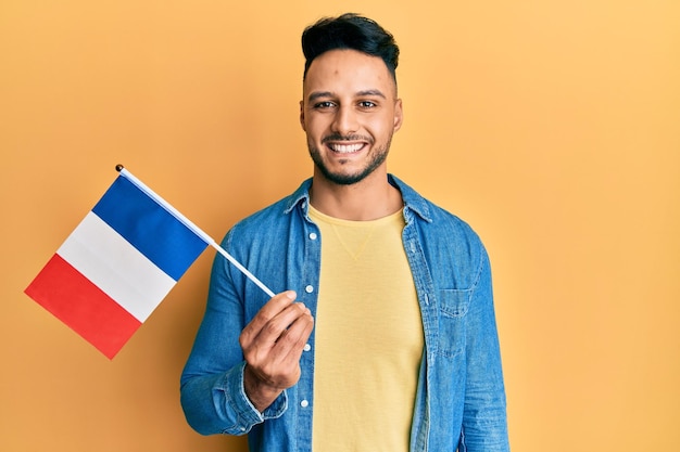 Gratis Foto jonge arabische man met de vlag van frankrijk die er positief en gelukkig uitziet en glimlacht met een zelfverzekerde glimlach die tanden laat zien