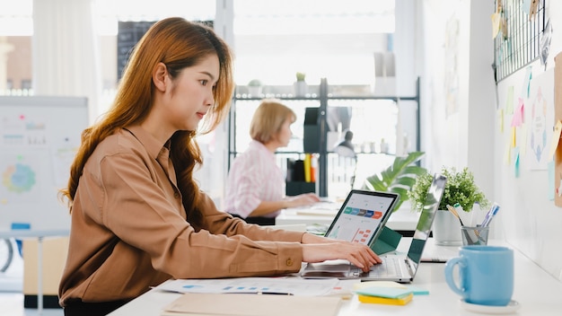 Gratis Foto jonge aziatische zakenvrouw ondernemer sociale afstand in nieuwe normale situatie voor viruspreventie tijdens het gebruik van laptopcomputer en tablet terug op het werk op kantoor.