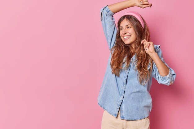 vrouw danst met opgeheven armen glimlacht breed beweegt actief draagt hoofdband en denim shirt geïsoleerd op roze kopie ruimte aan de linkerkant voor uw promotionele inhoud