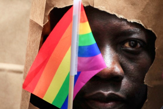 A man covers his face with a paper bag and holds a pride flag in front of his eye