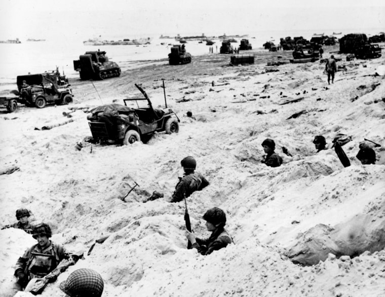 Sitting in the cover of their foxholes, American soldiers of the Allied Expeditionary Force secure a beachhead during the initial Normandy landing operations. In the background, amphibious tanks and other equipment crowd the beach, while landing craft bring more troops and material ashore.