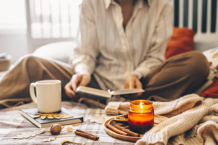 Women sitting on bed reading book. Mug coffee or tea and wooden tray with burning candle. Home decor elements.