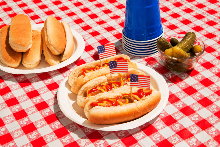 Photo of hot dogs with ketchup, mustard, and pickles on a checkered tablecloth. The hot dogs are decorated with patriotic American flag toothpicks. Paper plates and disposable plastic cups are also visible. The scene is outdoors on a sunny day, representing a carefree summer BBQ for Independence Day or Memorial Day. No people are in the photo.