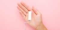 White sanitary tampon on young adult woman palm on light pink table background. Pastel color. Closeup. Female hygiene. Top down view.
