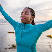 Celebration of Life. Beautiful woman if mixed race. Her arms raised up in the air. Her head is tilted back and eyes are close. She is in awe and smiling at the beauty of nature. Woman is expressing positivity, and feels grateful for life. She is very spiritual. Woman is at the beach with beautiful sunset in the background. 