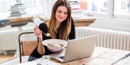 Young woman in city apartment eating muesli breakfast whilst reading laptop