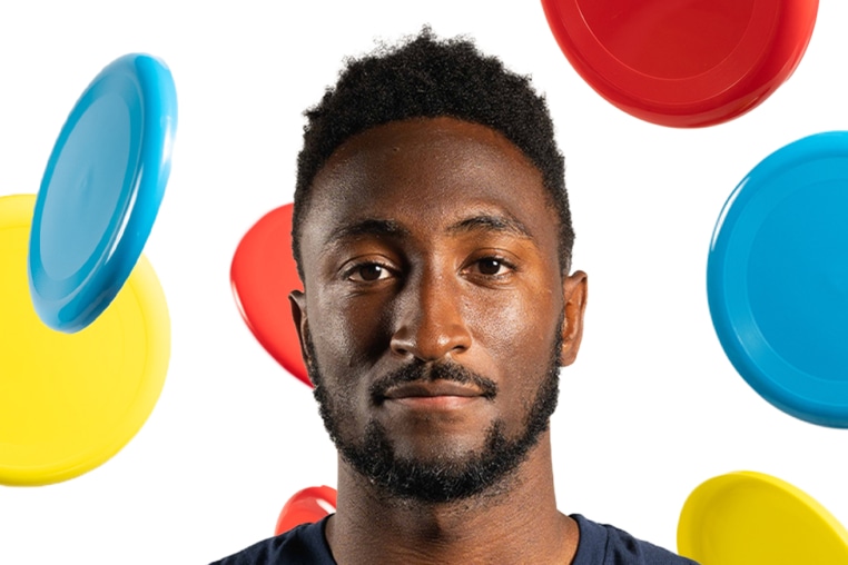 Marques Brownlee with his arms crossed with multicolored frisbees flying behind him.